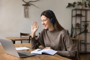 Cheerful freelance professional woman in big headphones speaking on video conference call, waving...
