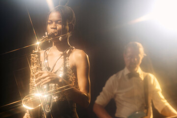 Portrait of Black young woman playing saxophone with jazz band on stage in hazy nightclub with golden lens flare accents copy space - obrazy, fototapety, plakaty