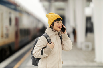 A happy middle-aged asian woman is standing at train station and talking on the phone.