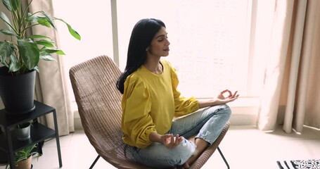 Serene Indian woman do meditation practice seated cross-legged in armchair, exhaling inhaling deeply, breath fresh conditioned air, relax indoor. Stress-free, boost mindfulness, yoga training at home