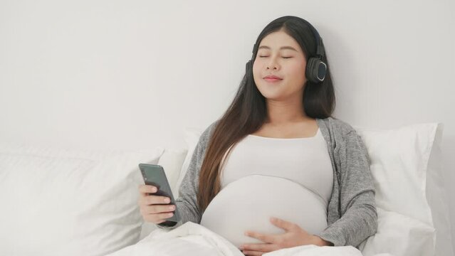 A pregnant woman wears headphones and listens to lullabies on the bed in the bedroom.