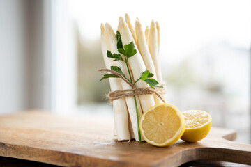 Standing bunch of fresh white asparagus. Seasonal spring vegetables with parsley and lemon on wooden cutting board. Kitchen scene for the seasonal gastronomy. - obrazy, fototapety, plakaty