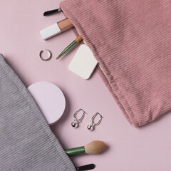 A Gray and Pink makeup cosmetic bag with cosmetic beauty tools and product spilling out on to a pastel colored pink background Top View Earrings Square