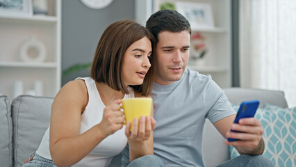Beautiful couple sitting on sofa together using smartphone drinking coffee at home