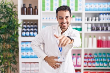 Handsome hispanic man working at pharmacy drugstore laughing at you, pointing finger to the camera with hand over body, shame expression