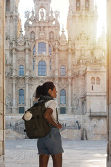 A woman carrying a backpack in which she carries a scallop shell as a symbol of the Camino de Santiago, arriving at the Cathedral of Santiago de Compostela. - obrazy, fototapety, plakaty