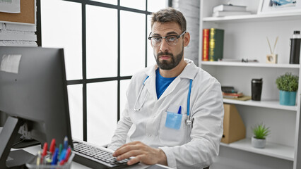 Bearded young hispanic man doctor hard at work, using his computer indoors at a medical clinic