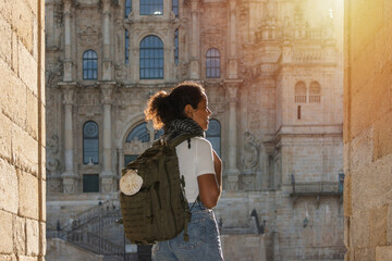 Fototapeta na wymiar An African pilgrim woman, seen from behind, carrying a backpack adorned with a scallop shell symbol, entering the square of Santiago Cathedral illuminated by the morning sun