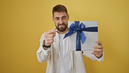 Cheerful young hispanic man points his finger at you, holding a birthday gift box isolated on a yellow background, smiling and showing confidence