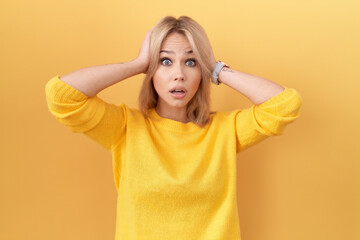 Young caucasian woman wearing yellow sweater crazy and scared with hands on head, afraid and surprised of shock with open mouth