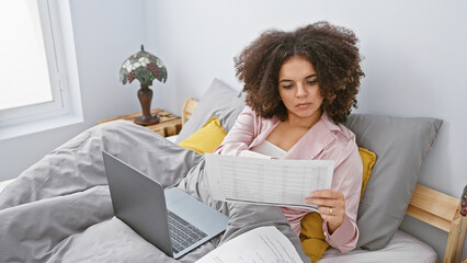 Hispanic woman with curly hair examines documents in a bedroom with a laptop nearby, conveying a...