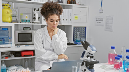 Hispanic woman working thoughtfully in a laboratory with microscope and laptop