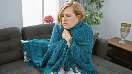 Naklejka premium Mature caucasian woman feeling unwell wrapped in a blanket sitting on her living room couch indoors.