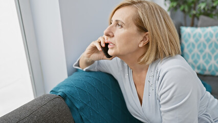 Mature blonde woman conversing on a smartphone in a cozy home interior, evoking a sense of everyday...