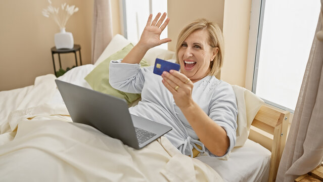 Smiling middle-aged woman with credit card waving at laptop in bedroom, depicting online shopping or virtual greeting.