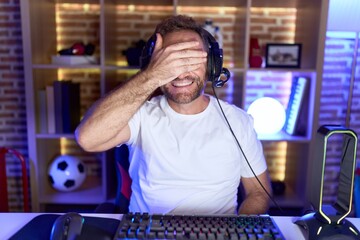 Middle age man with beard playing video games wearing headphones smiling and laughing with hand on face covering eyes for surprise. blind concept.