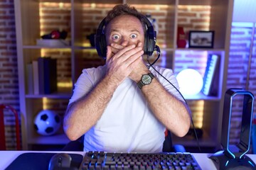 Middle age man with beard playing video games wearing headphones shocked covering mouth with hands for mistake. secret concept.