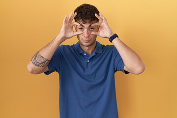 Young hispanic man standing over yellow background trying to open eyes with fingers, sleepy and tired for morning fatigue