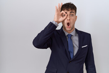 Young hispanic business man wearing suit and tie doing ok gesture shocked with surprised face, eye looking through fingers. unbelieving expression.