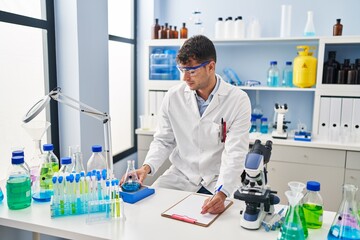 Young hispanic man scientist weighing liquid writing on document at laboratory