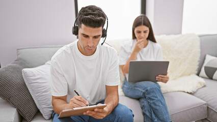 A man with headphones focused on writing while a woman works on a laptop in a modern living room.