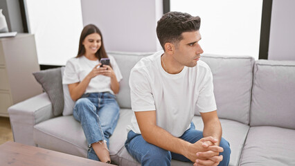 A man and woman in casual clothing sit separately on a gray couch in a modern living room, with the woman smiling at her smartphone.