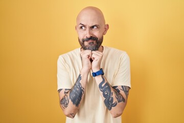 Hispanic man with tattoos standing over yellow background laughing nervous and excited with hands...