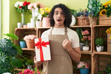 Hispanic man with curly hair working at florist shop holding gift scared and amazed with open mouth for surprise, disbelief face