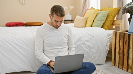 A young hispanic man with a beard works on a laptop in a cozy bedroom setting.