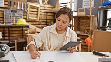Attractive young hispanic woman carpenter immersed in her woodworking craft, drawing on a touchpad at carpentry workshop