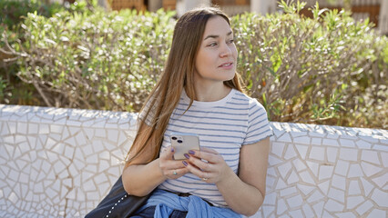 A contemplative young woman with long hair holds a smartphone in a sunny park, emanating serene beauty amidst nature.