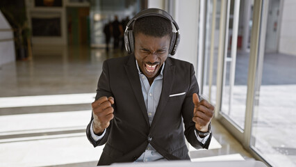 Excited african american man in business attire with headphones celebrating in a modern office...
