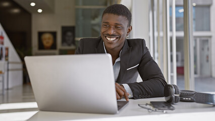 A cheerful african man works on a laptop in a modern office setting, portraying business,...