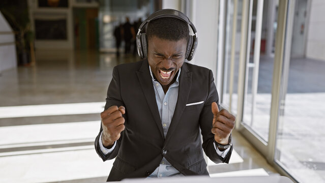 Excited african american man in business attire with headphones celebrating in a modern office setting.