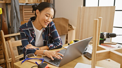 Smiling asian woman using laptop in a woodworking workshop surrounded by tools and timber.