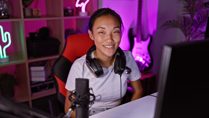 A smiling young asian woman with headphones in a colorful neon-lit gaming room at night. © Krakenimages.com
