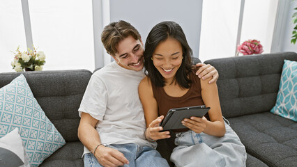 Happy interracial couple sharing a moment with tablet on cozy sofa indoors, expressing affection...