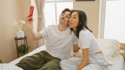 Obraz premium An interracial couple takes a playful selfie in a well-lit bedroom, showing affection and togetherness in a cozy home setting