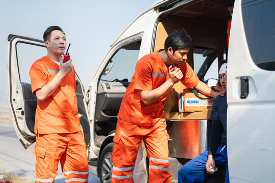 Asian male EMS paramedics or emergency medical technicians in orange uniforms performing first aid applying bandage onto head of young man patient in ambulance from road accident