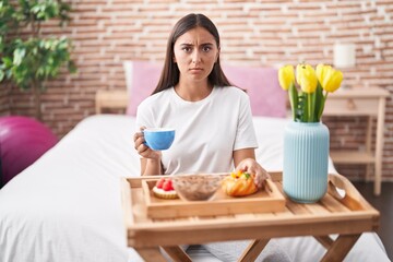 Young hispanic woman eating pastries for breakfast sitting on the bed skeptic and nervous, frowning...