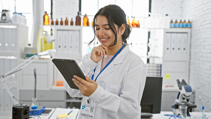 A smiling young hispanic woman in a lab coat uses a tablet in a modern laboratory, representing...
