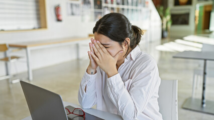 A stressed hispanic woman in a white shirt covers her face with her hands at a modern office table, with a laptop and glasses nearby.