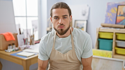 Young hispanic man artist sitting on chair with serious face at art studio