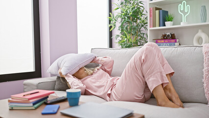 Young blonde woman lying on sofa covering face with cushion at home