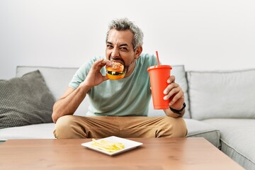 Middle age grey-haired man eating hamburger sitting on sofa at home