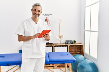 Middle age grey-haired man physiotherapist smiling confident using touchpad at rehab clinic