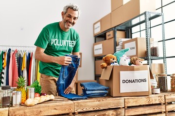 Middle age grey-haired man volunteer smiling confident folding jeans at charity center