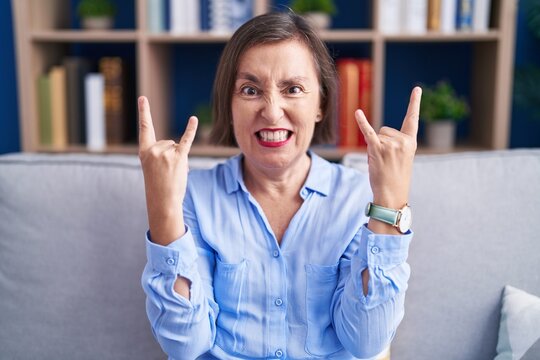 Middle age hispanic woman sitting on the sofa at home shouting with crazy expression doing rock symbol with hands up. music star. heavy concept.