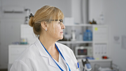 Focused middle age blonde woman scientist, serious expression, standing side-face in lab, immersed...
