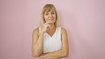 Middle age beautiful blonde woman wears serious expression, standing confidently over isolated pink background, deep in thought